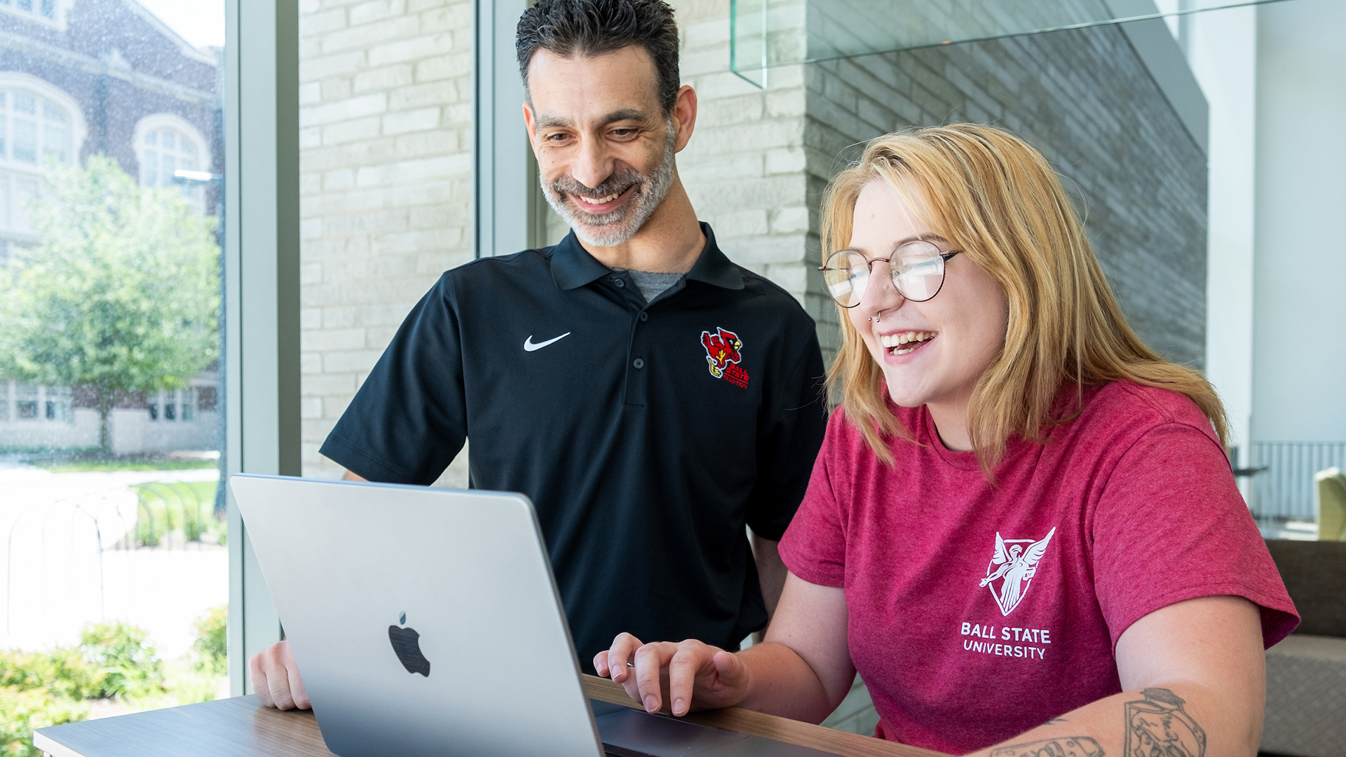 A girl wearing glasses and a red shirt and a man wearing a black polo sit next to each other in a naturally-lit room while they smile at a laptop screen