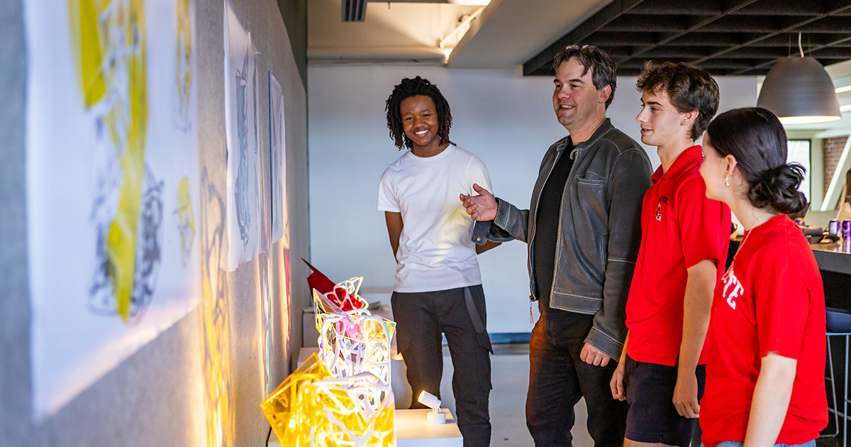 Three students and a faculty member smile and look at architectural renderings on a gallery wall
