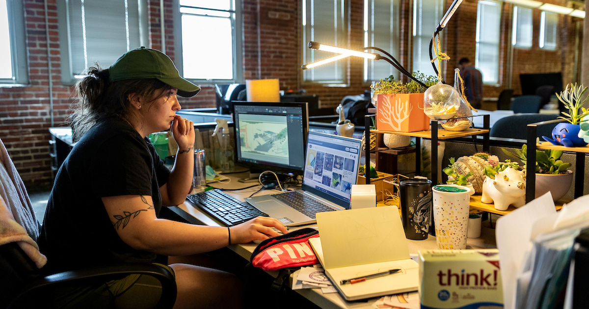 Architecture students looks at a laptop screen while sitting at a desk that has several small plants on it 
