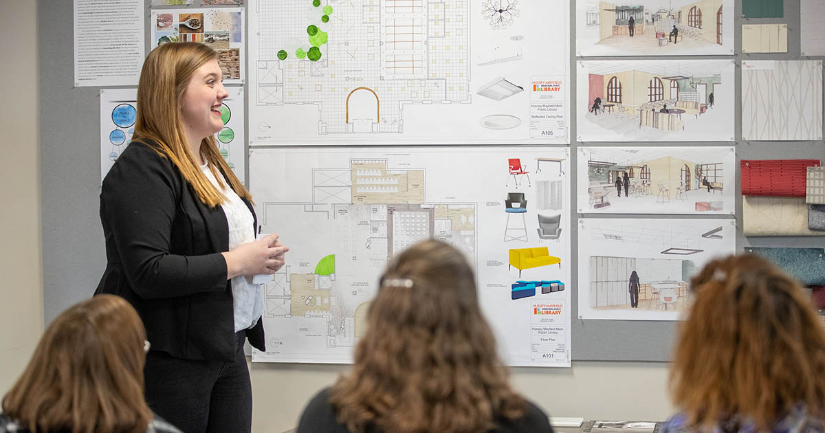 A young woman wearing a black pantsuit stands in front of a classroom and looks at a board displaying various architectural renderings. 