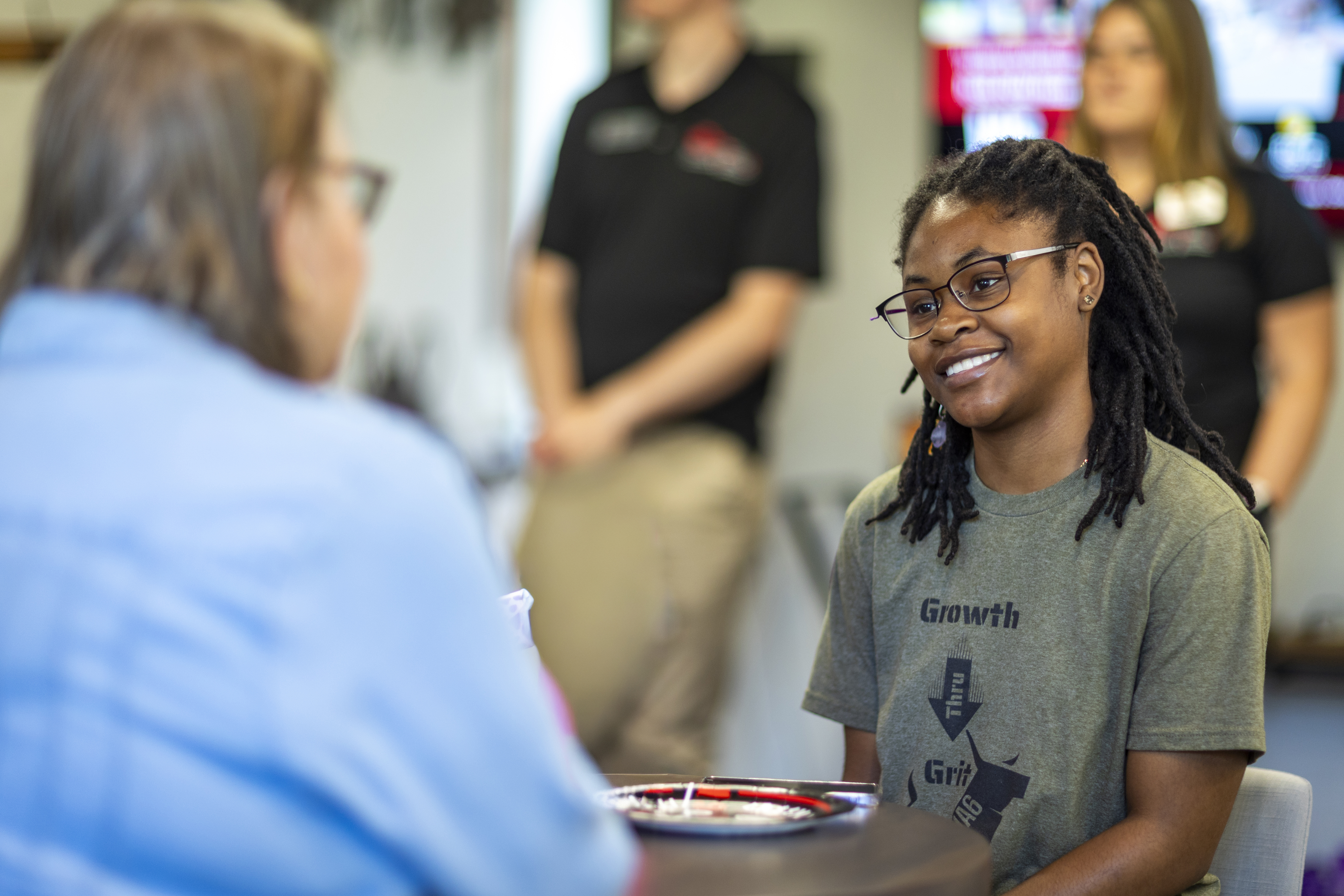 Student in the connection lounge with dark complexion dark medium long hair with glasses and a green shirt sitting across from a networking partner in a blue shirt only see the back of the person in blue shirt