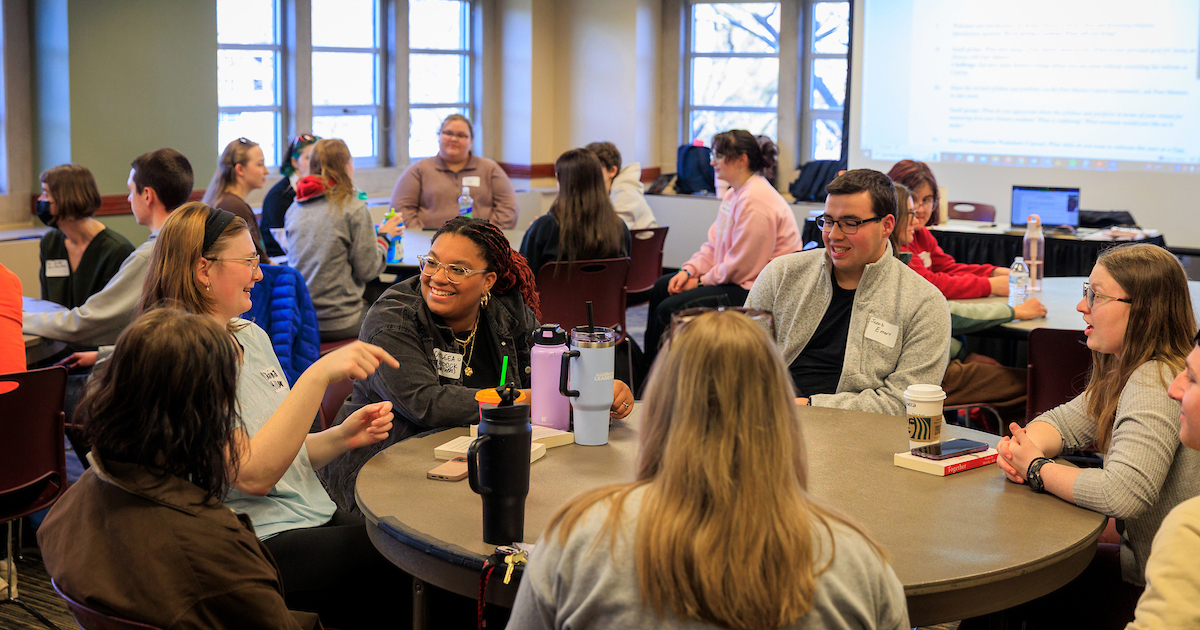 Students sit around tables in a classroom, talking and collaborating during a group discussion.