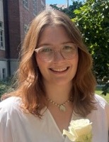 Headshot of a smiling woman with medium-length, reddish-brown hair wearing a white blouse and glasses while holding a white rose