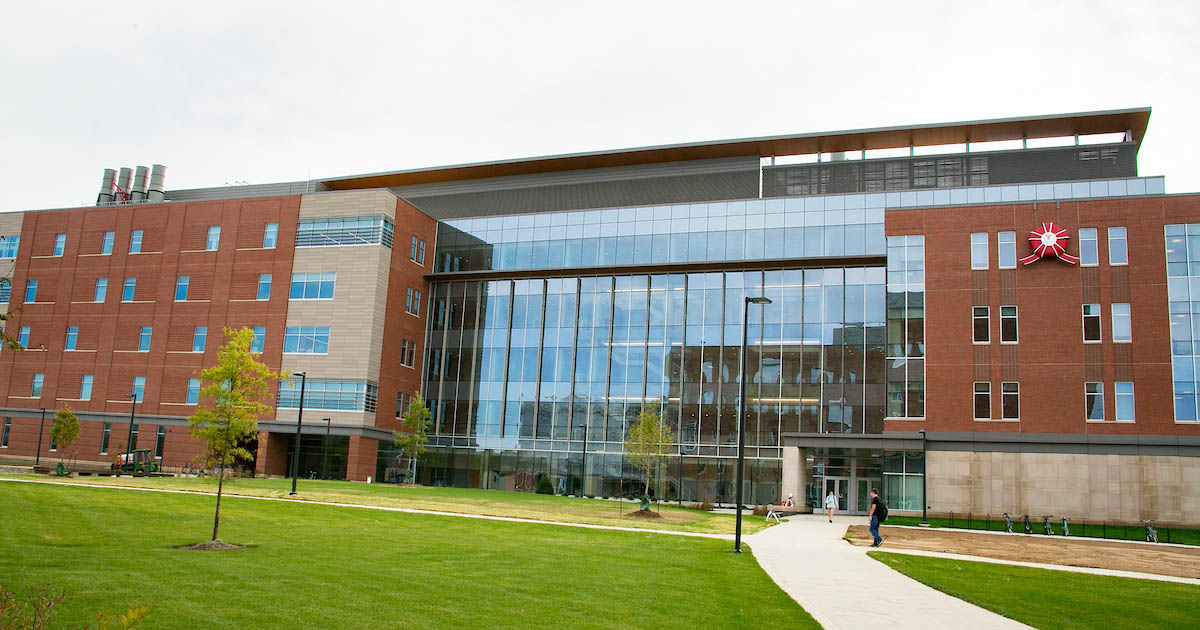 Ball State's Foundational Sciences Building, a modern building with red brick and various windows underneath a cloudy sky and surrounded by green grass.