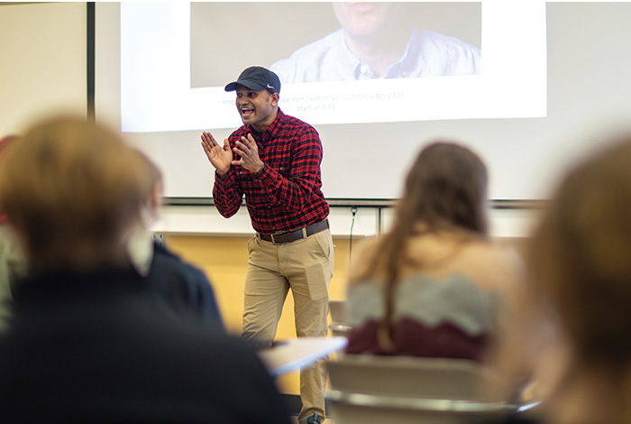 Student with red shirt and ball cap speaks in front of classroom of students