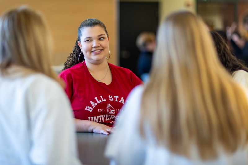 A student wearing a Ball State University T-shirt smiles while talking with a small group of peers gathered around a table, with two participants blurred in the foreground.