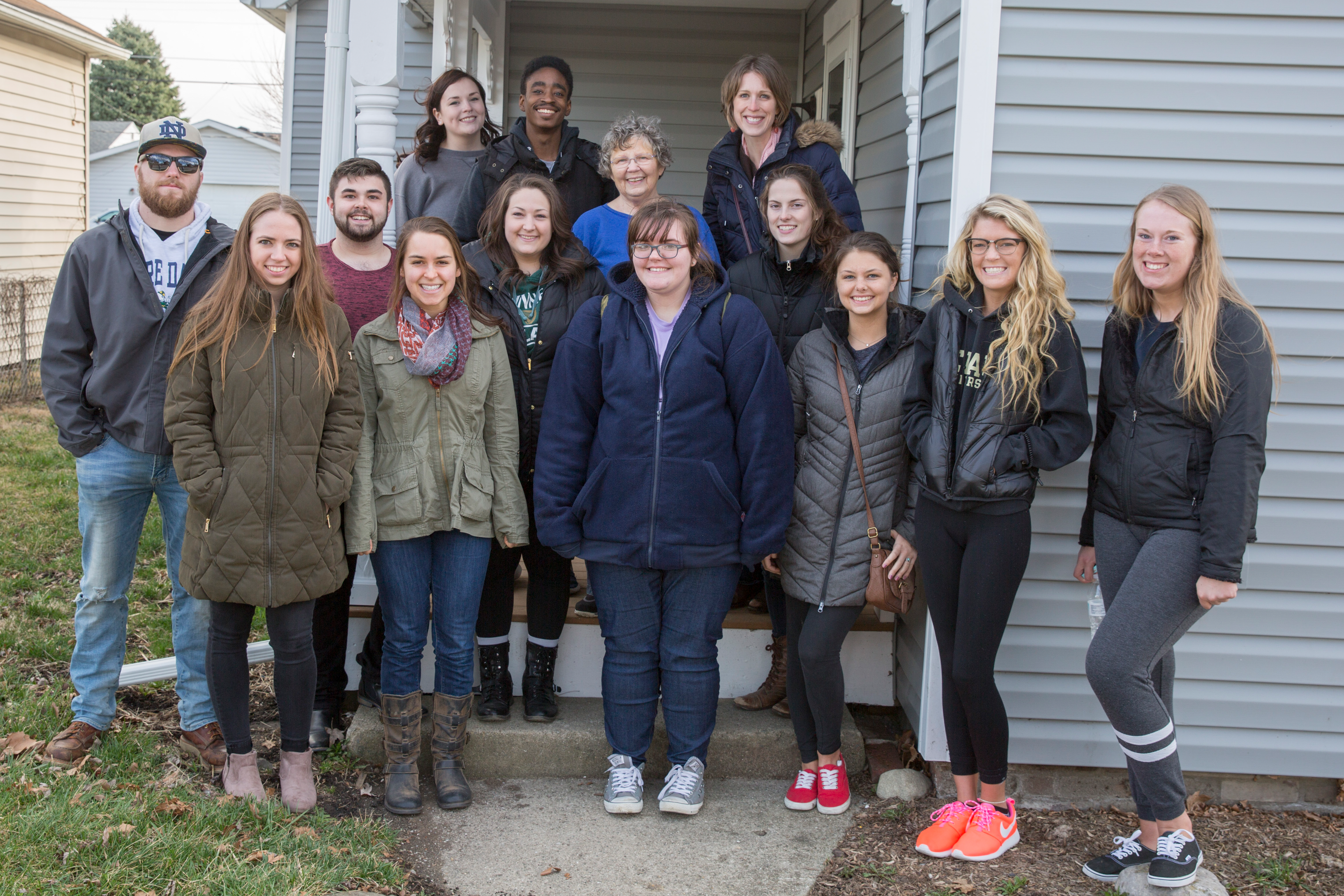 A group of students and project members poses for a group photo in front of a blue house with white trim
