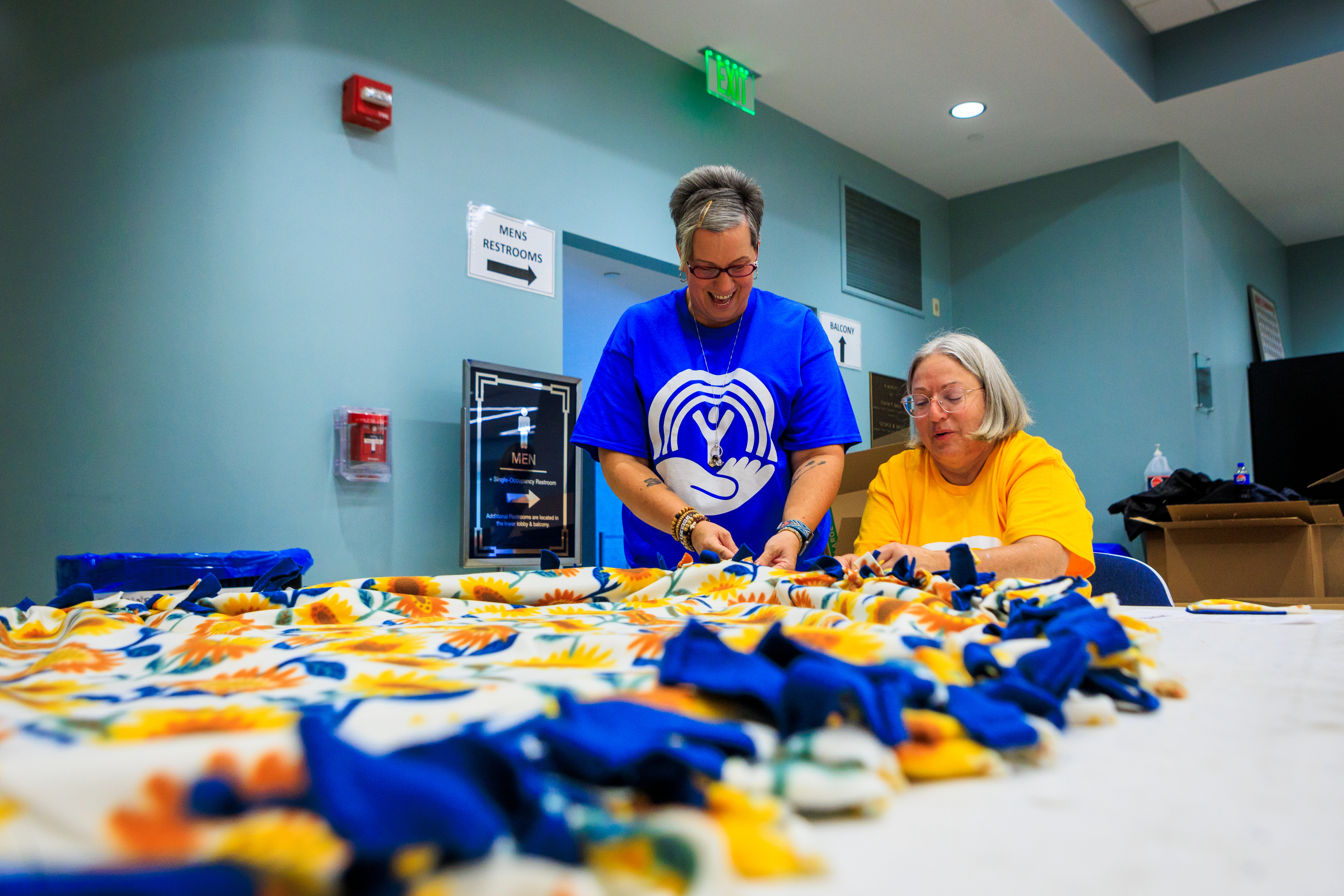 A woman in a blue volunteer t-shirt and a woman in a yellow volunteer t-shirt sit at the end of a table. In the foreground and stretching toward the employees is a blue and yellow felt blanket they are knotting for the unhoused.