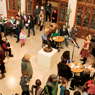 Many people mill around on the white marble floor of the museum lobby while a woman in a red dress sings at a piano and people do crafts off to the sides.