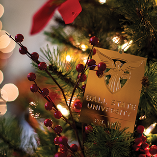 A rectangular golden holiday ornament in a dark green, red and gold decorated tree. The Ball State logo is etched into the ornament