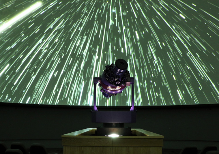 The planetarium projector is silloutetted against a background of stars streaming through a green sky