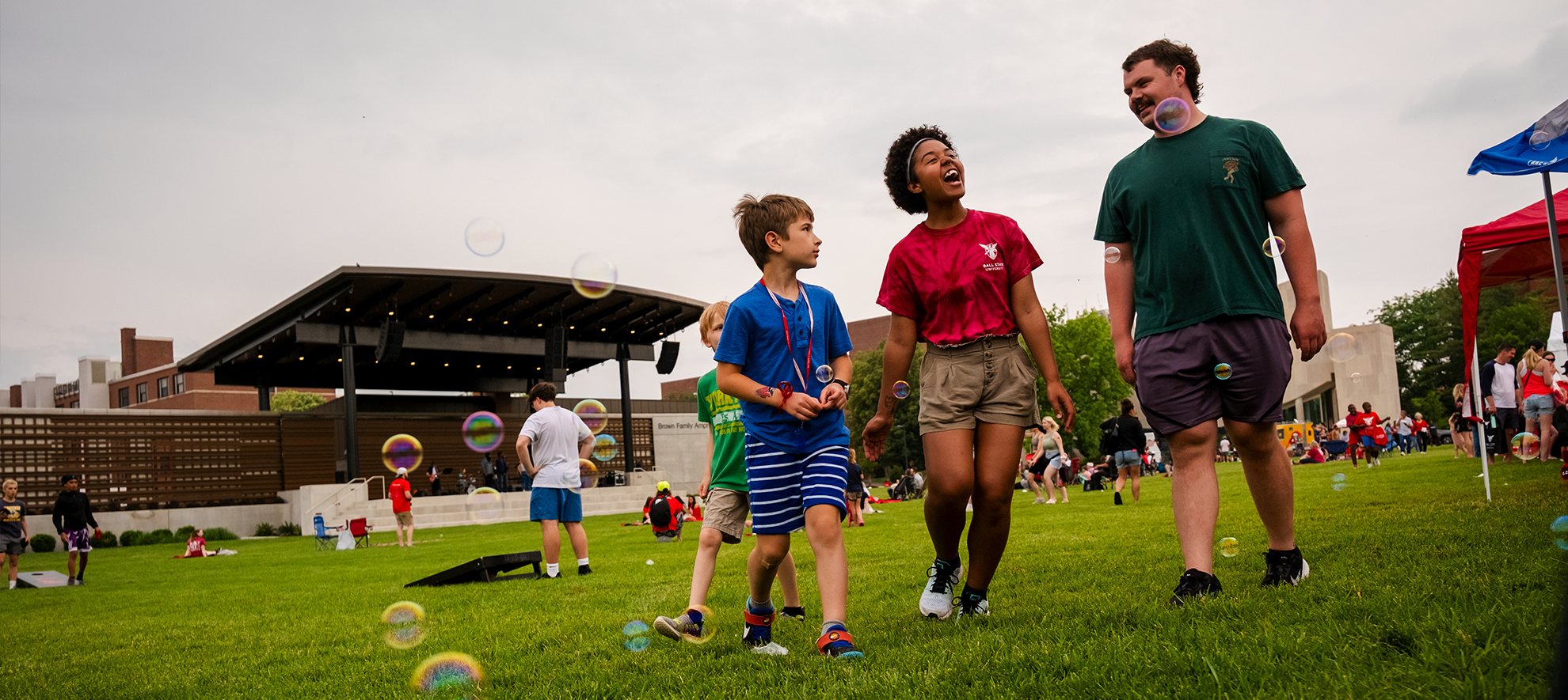 Groups of people of all ages and backgrounds enjoy games on the amphitheater lawn while a child and two adults walk laughing toward the camera.