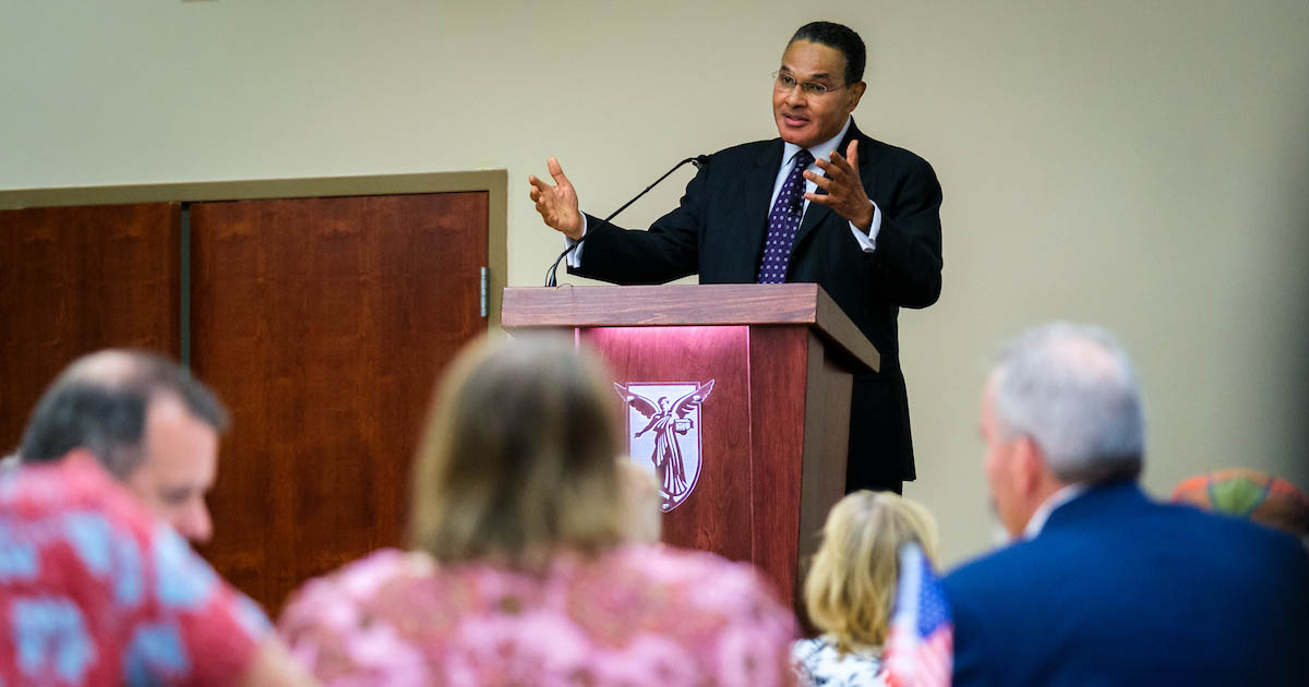 A man wearing a black suit and blue, dotted tie stands in front of a room of people at a podium while gesturing with both hands outwardly