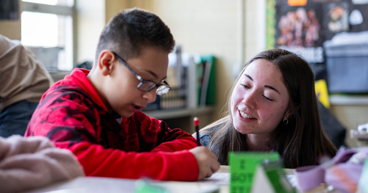 A young woman with long, dark hair smiles and crouches down to assist a young student who is working on an assignment at a desk