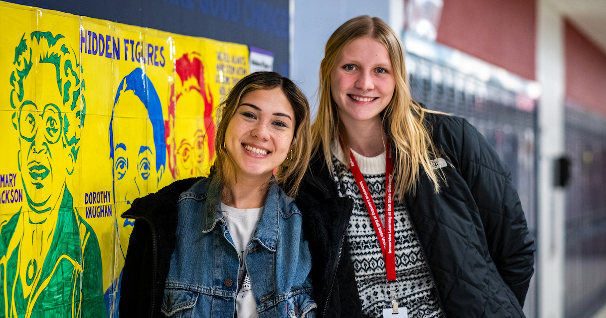 Two young woman pose and smile next to each other while standing in a school hallway with lockers and posters in the background.