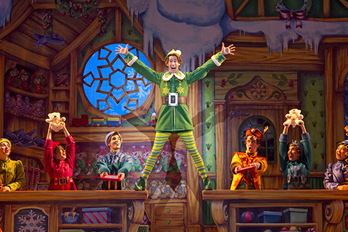 Buddy the Elf standing on two tables in a toy shop with other elves. 