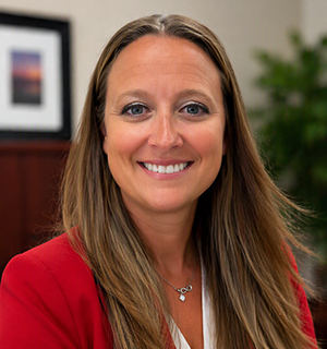 Carrie smiles for a professional headshot wearing a red blazer in an office setting.