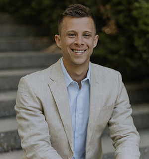 Roman B. smiles for an outdoor professional portrait wearing a light-colored blazer.
