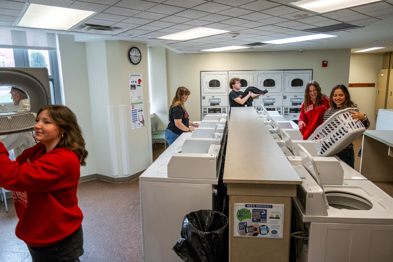 Students in a laundry facility with washers and dryers and holding a basket doing laundry