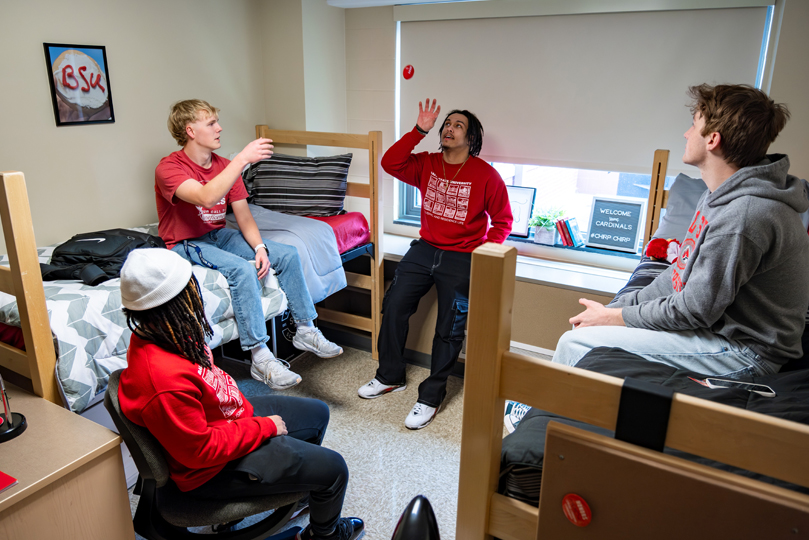 Four students in a residence hall room. One sitting down in a chair, one on the bed to the left, one sitting near the window and one sitting on the bed to the right