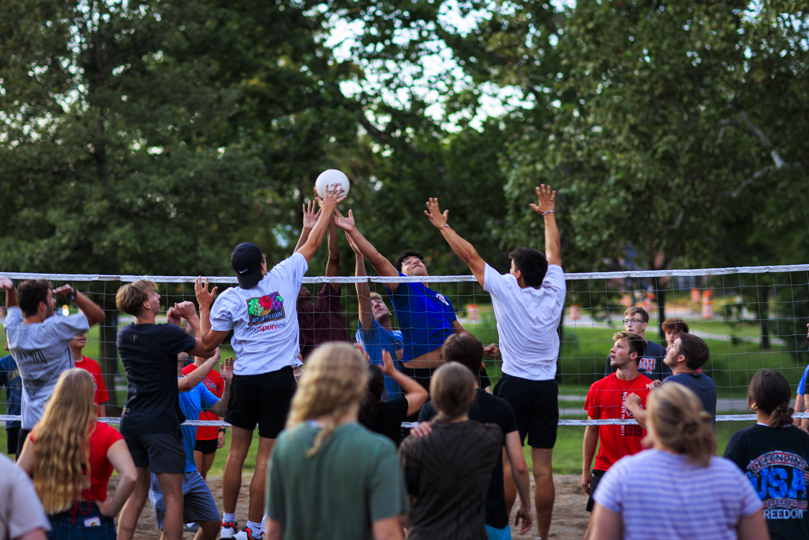 large group of students outside at the volleyball courts, students jumping up near the net to hit the volleyball 