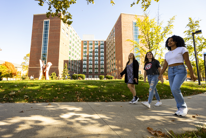 three female students walking outside on the sidewalk with a residence hall in the background 