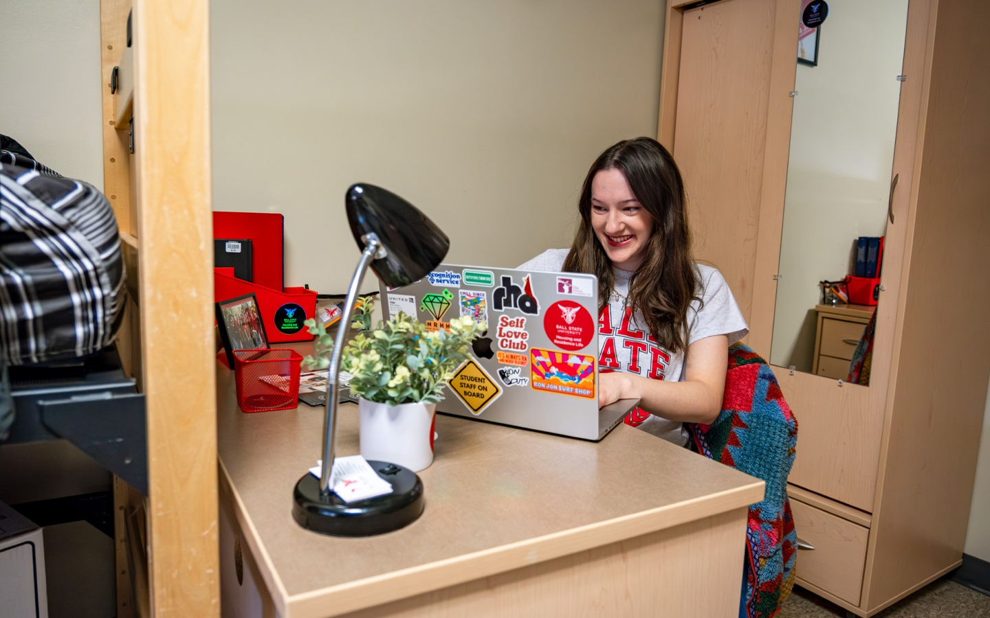 female student sitting at desk with a laptop, a light on the desk and a dresser/wardrobe in the background