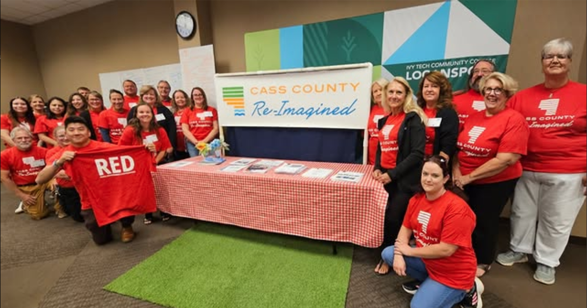 Group in matching red shirts poses at a Cass County Re-Imagined booth during a community event.