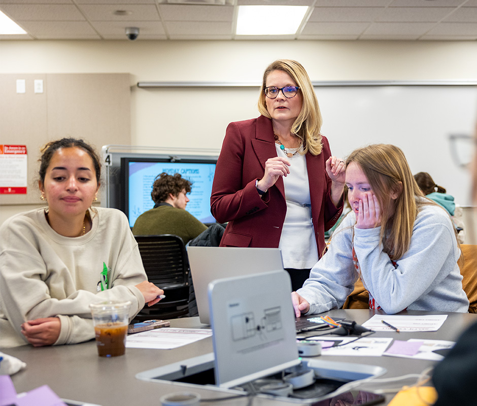 Two students at a desk on looks at a computer the other reading paper with teacher behind them