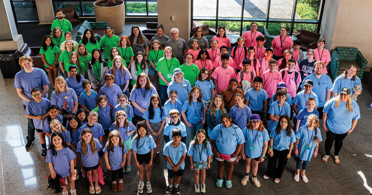 Group of children, teens, and adult leaders wearing color-coded camp T-shirts pose together in a large indoor lobby.