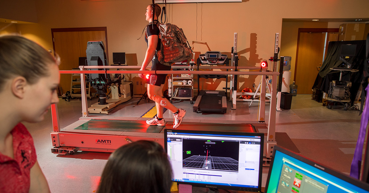 Two students look-on as a man runs on a treadmill.