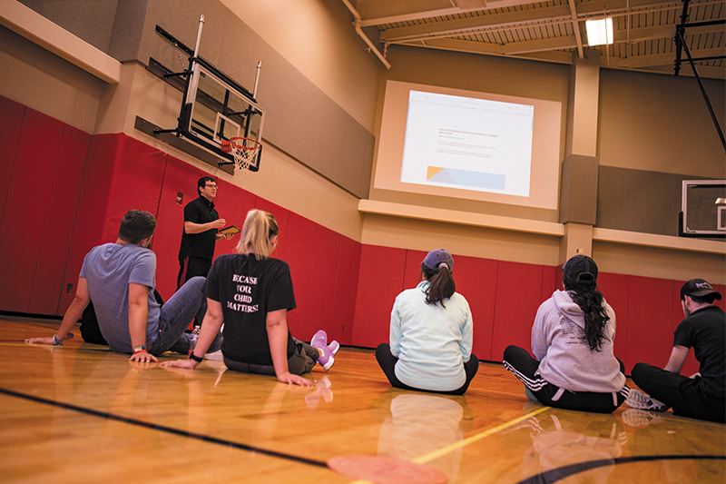 Students sit on gym floor listening to coach.