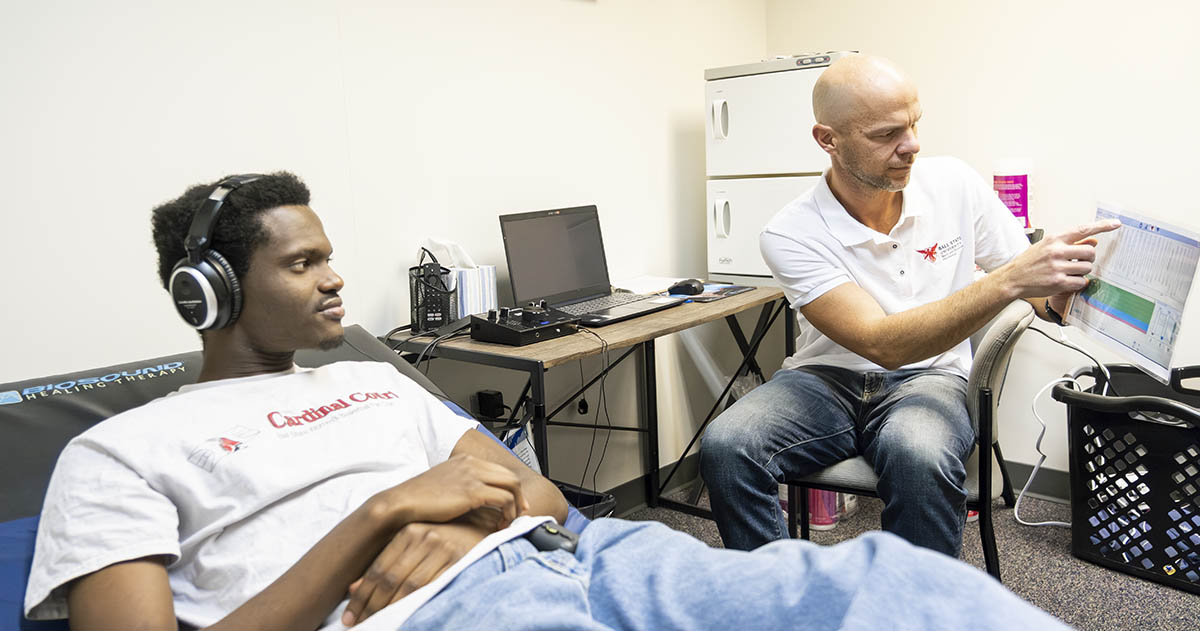 Two Sports Psychology students look at a sheet with lab work on it. One student is laying on a padded table wearing headphones while the other one sits in a chair and points at the lab work. 