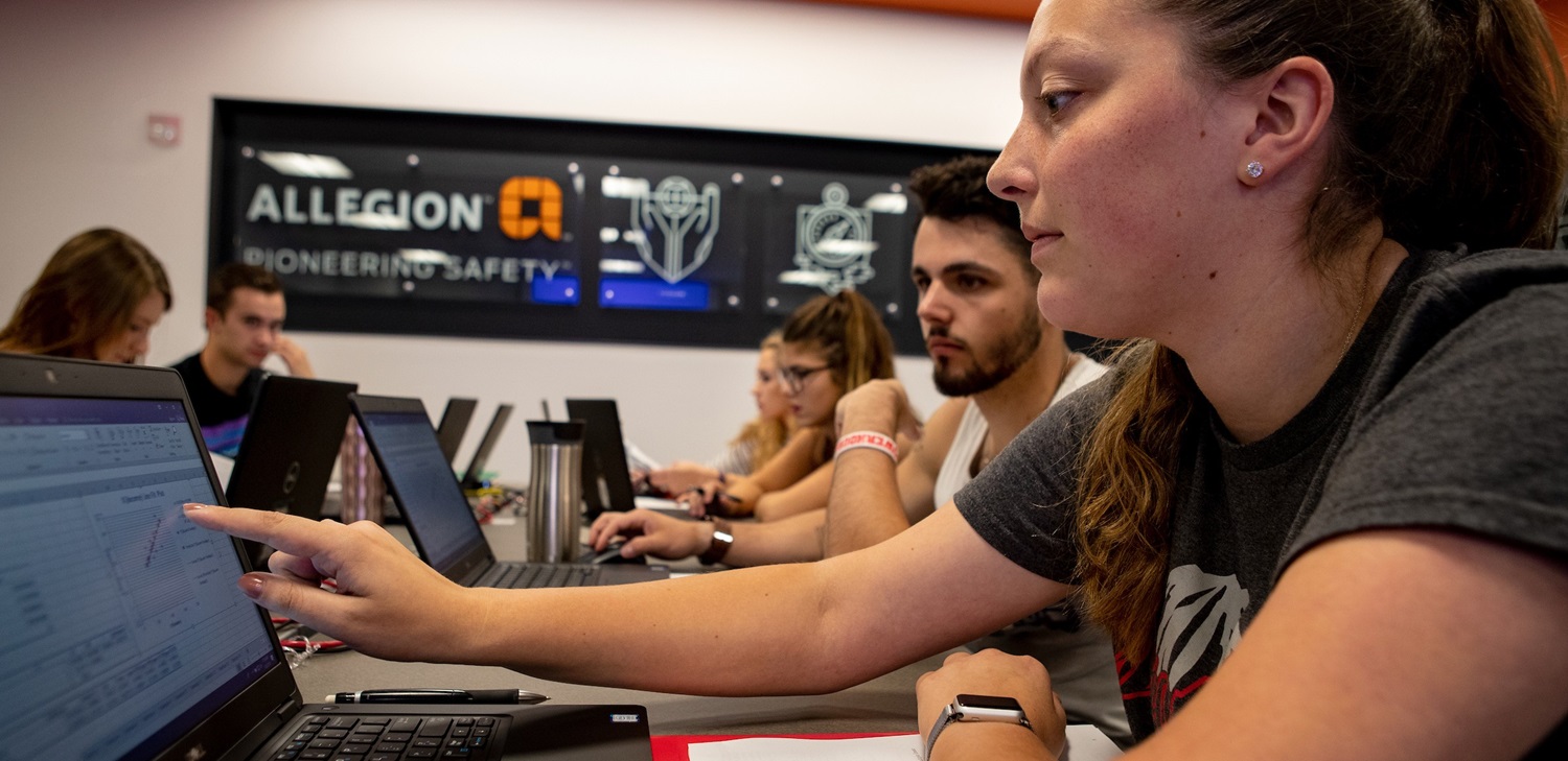 A woman in a grey t-shirt points at a data chart on her laptop screen while sitting at a long table with colleagues. In the background, other individuals work on their computers in front of a wall featuring the Allegion "Pioneering Safety" logo.
