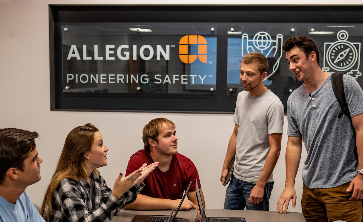 Five young adults gather around a conference table with open laptops, talking and smiling during a collaborative meeting. A wall sign behind them reads “Allegion – Pioneering Safety,” suggesting a workplace or corporate training setting.