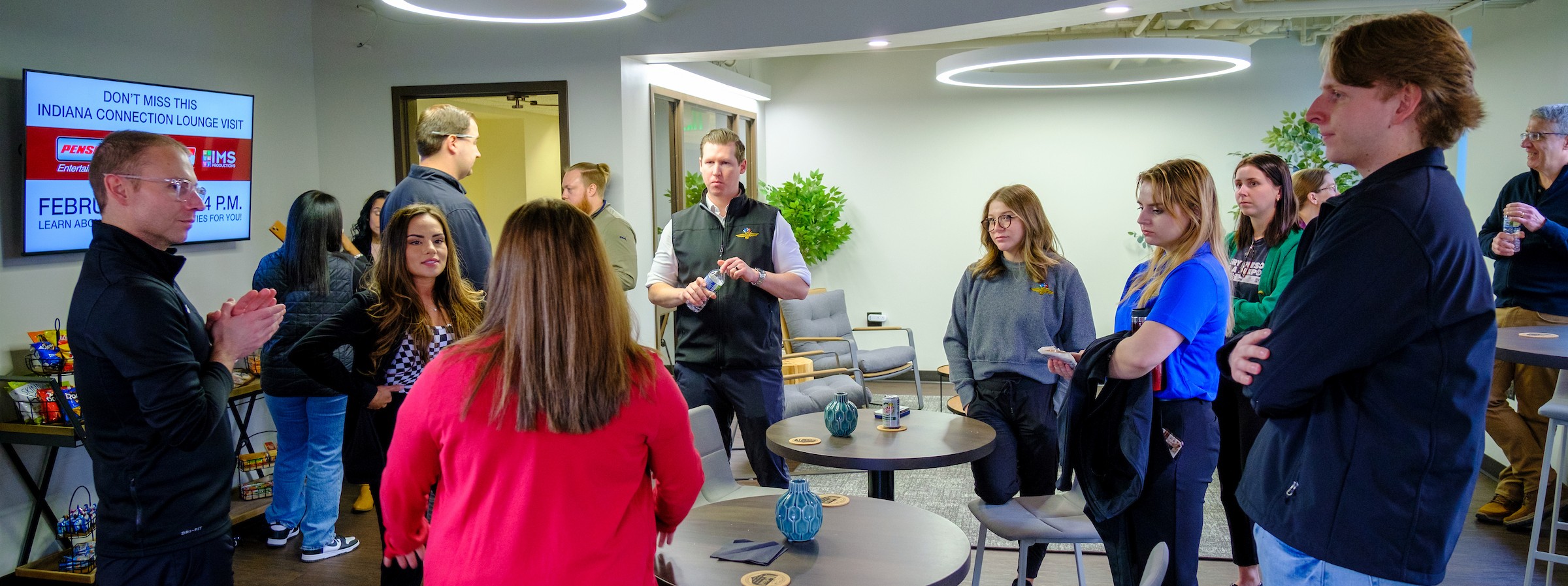 Gemini said A group of students and professionals stand and talk in a modern lounge area featuring circular overhead lights and small round tables. A television screen on the left wall displays an announcement for an "Indiana Connection Lounge Visit" featuring the Penske and Indianapolis Motor Speedway logos.