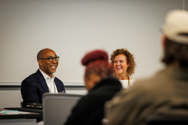  Gemini said In a bright classroom setting, a man in a navy blazer and glasses and a woman with curly blonde hair sit at the front of a room, both laughing during a presentation or discussion. The shot is taken from behind an audience, with the blurred figures of other attendees in the foreground.