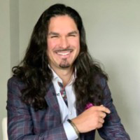 A portrait of Sahar Montalvo with long, wavy dark hair and a goatee, smiling broadly. He is wearing a patterned navy and purple plaid blazer over a light-colored shirt with a decorative collar. He is posed with one hand slightly raised, set against a plain, light-colored background.