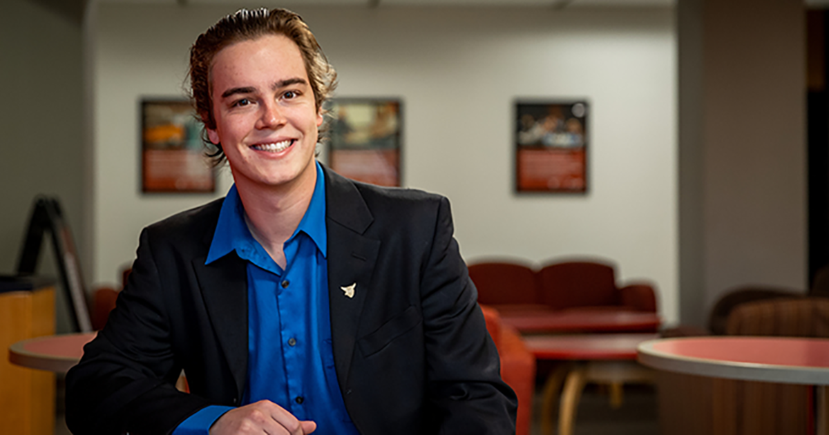 A young man with short blond hair smiles and poses while sitting at a table with other tables visible in the background, wearing a black blazer and bright blue undershirt