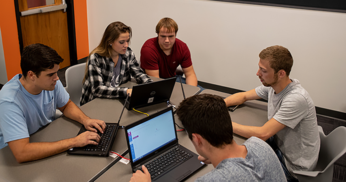 a group of five student sit at a classroom table facing each other with their laptops