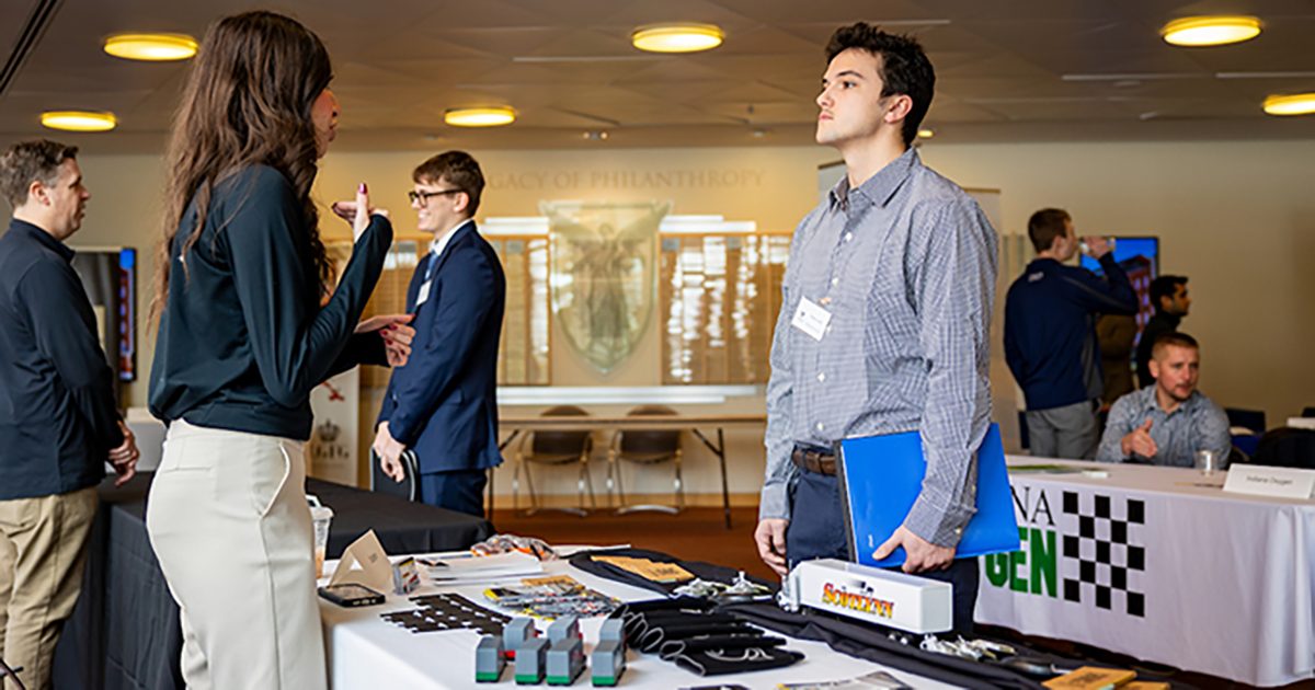 A student dressed in a long-sleeved button up shirt speaks with a woman at an event table