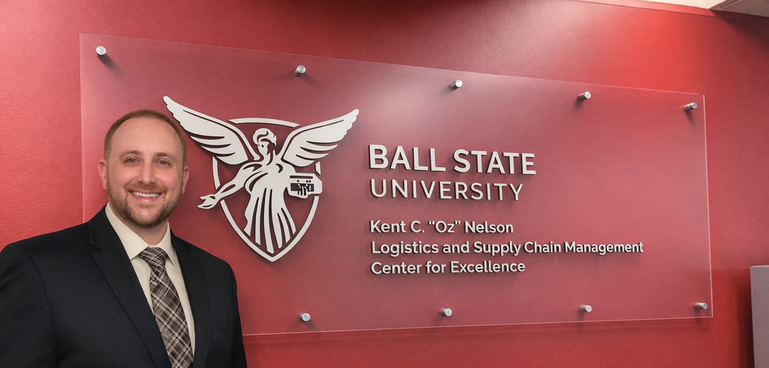  Gemini said Michael Gambrel, Director of the Logistics and Supply Chain Center for Excellence, stands next to the Ball State University Kent C. "Oz" Nelson Center signage on a red wall.