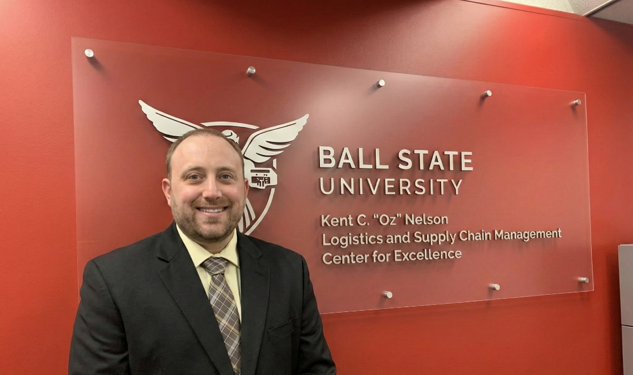 A man with short hair and a beard, wearing a black suit jacket, a light yellow shirt, and a plaid tie, smiles in front of a red wall. Mounted on the wall is a frosted glass sign with the Ball State University logo and text that reads: "BALL STATE UNIVERSITY Kent C. 'Oz' Nelson Logistics and Supply Chain Management Center for Excellence."