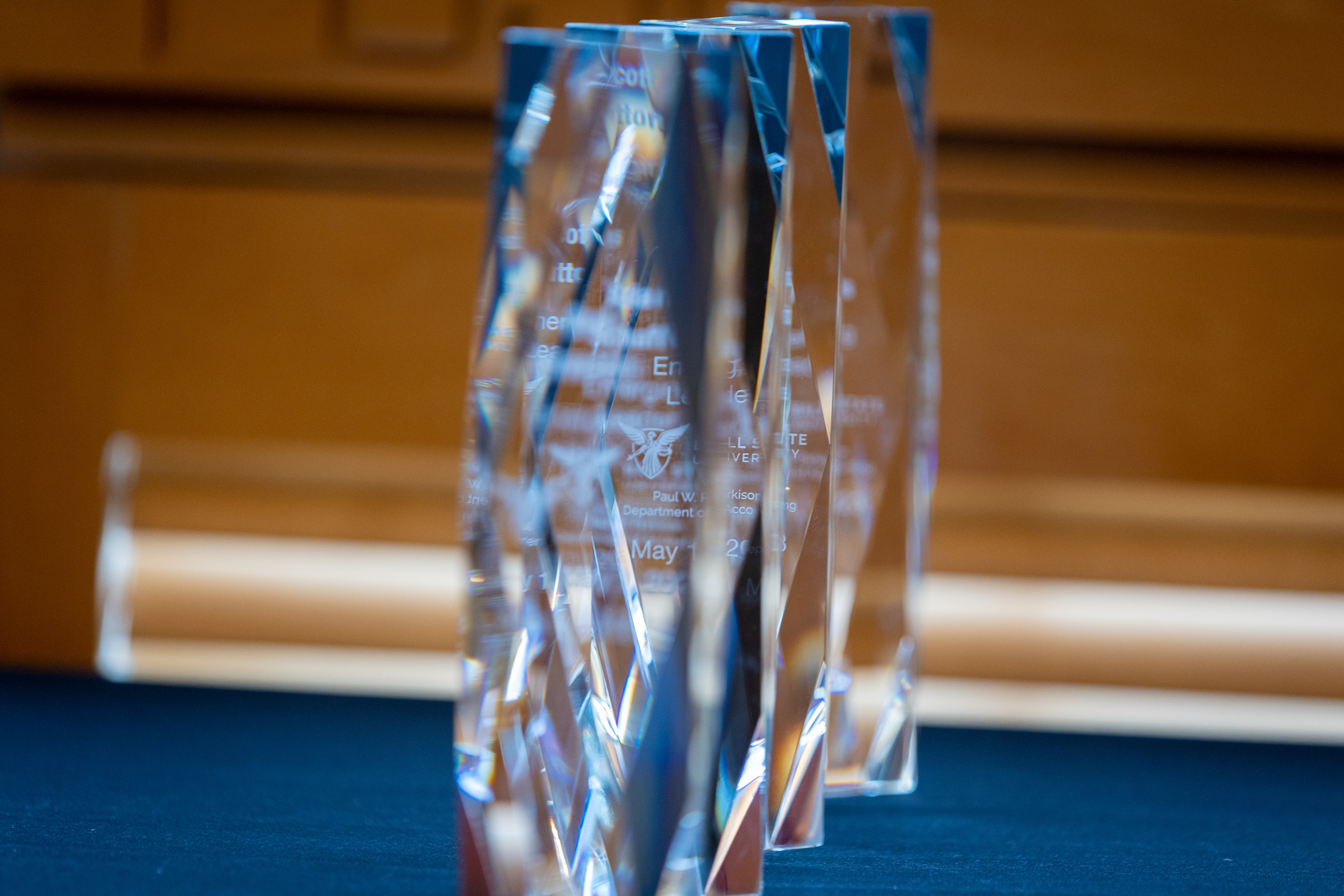 Several tall, clear glass or crystal awards with angular, faceted designs stand in a row on a dark blue tablecloth, with a softly blurred wood-paneled background behind them.