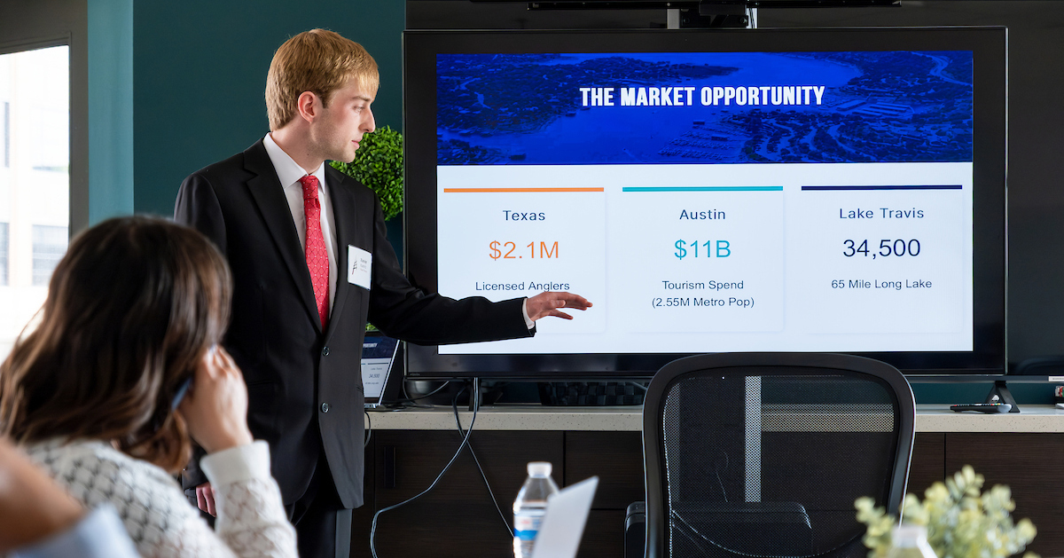 Student dressed in a suit and tie points at a screen displaying marketing statistics with attendees listening in a conference room