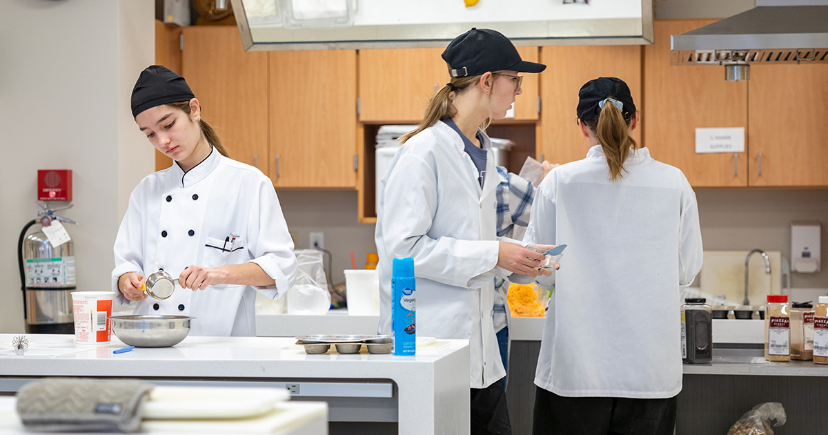 Students in chef coats prepare ingredients and recipes in a teaching kitchen as part of a culinary program.