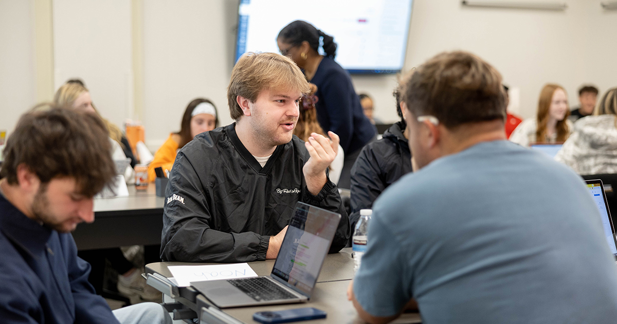 Students collaborate around a table during a classroom discussion, using laptops and sharing ideas in an active learning environment.