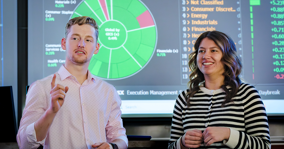 Students present financial market analysis in front of investment data and charts displayed on a classroom screen.