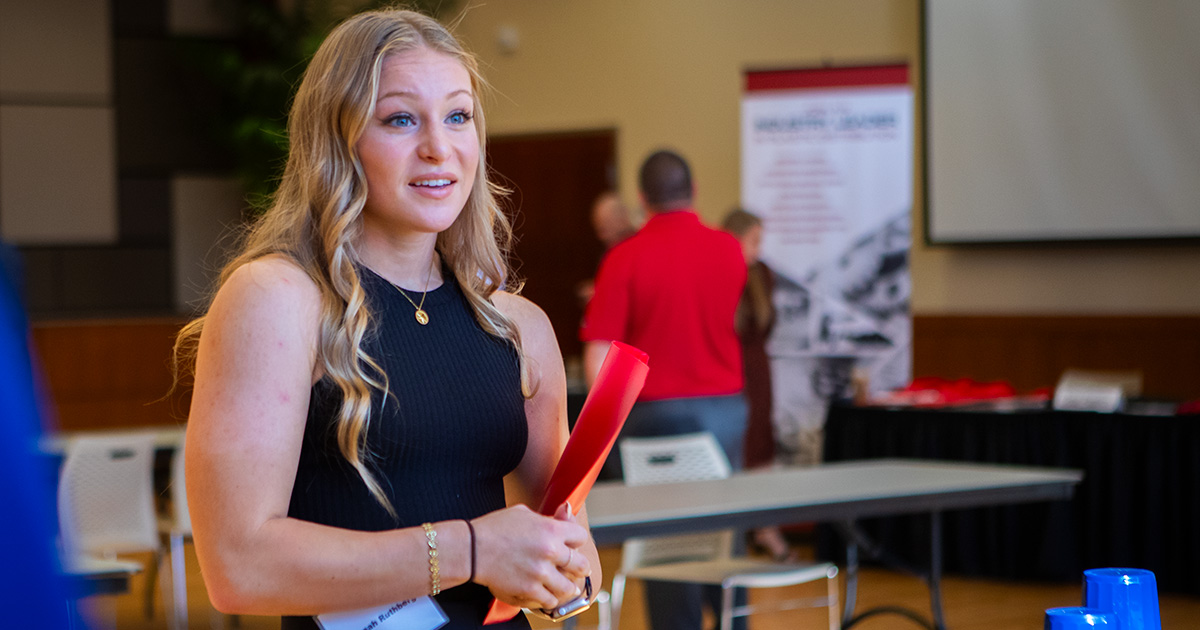 A girl with long blonde hair dressed in a sleeveless black top speaks to a professional at an event while holding a red folder