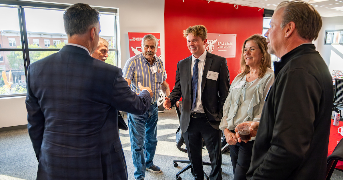 A group of professionally-dressed people smile and chat to each other in a well-lit room and red walls. Two of them shake hands across the group.